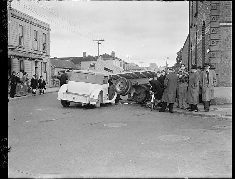 A crowd gathers to look at a collision. The accident took place in front of the Mount Cook Barracks and the Salvation Army at the intersection of Tasman and Buckle Streets in Wellington, 1950.